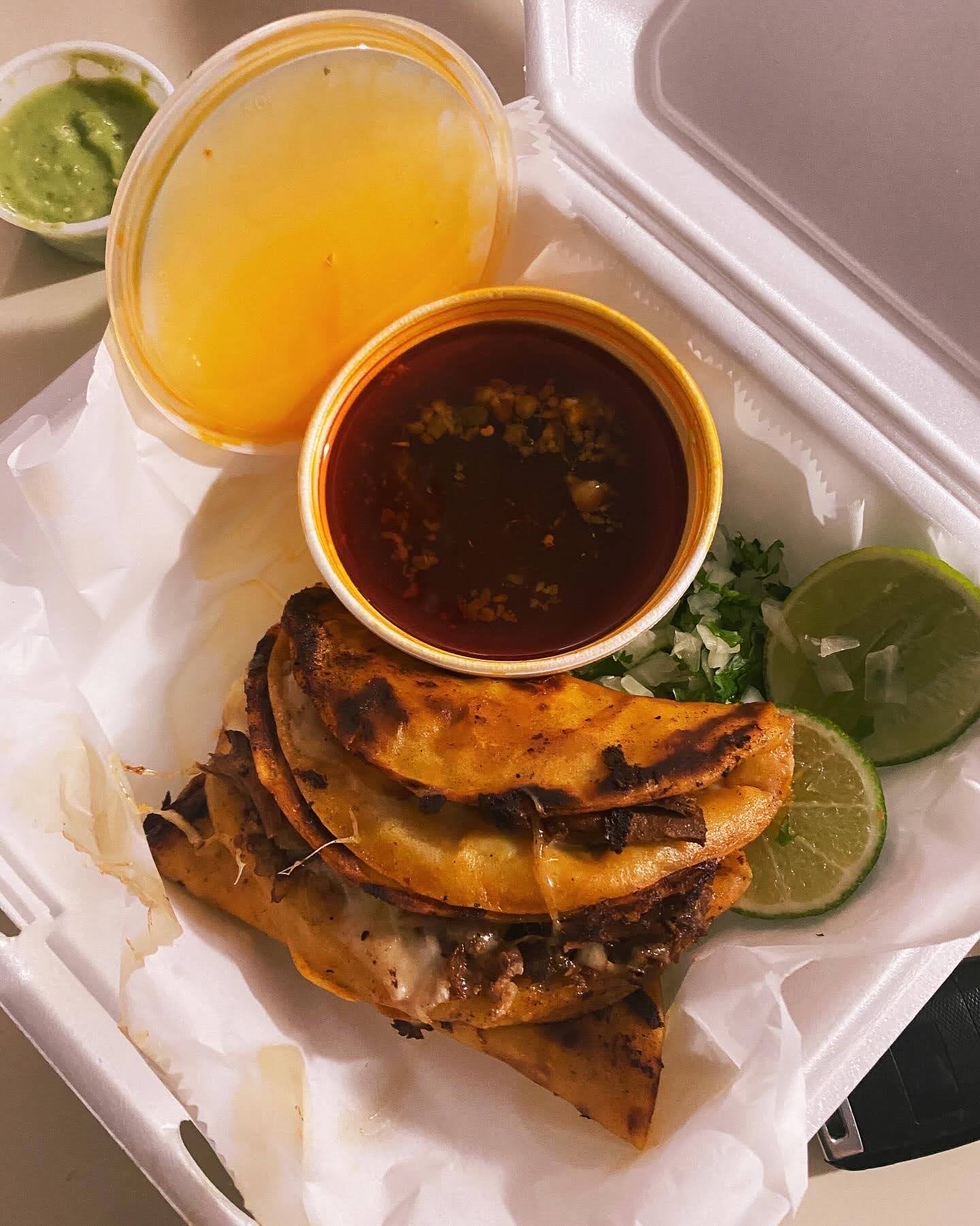 Birria tacos in a takeout box with a cup of consomé and a lime